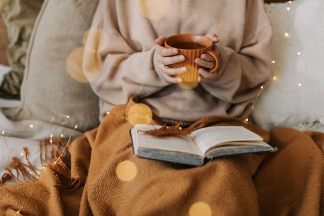 autumn indoor lifestyle image of person covered in blanket, reading a book and holding a cup